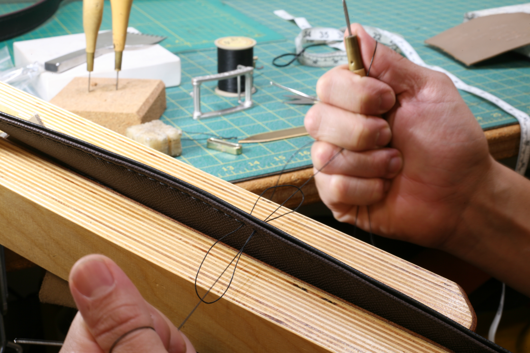 Close-up of a craftsman hand-stitching a black leather belt on a wooden clamp for men's premium belts.