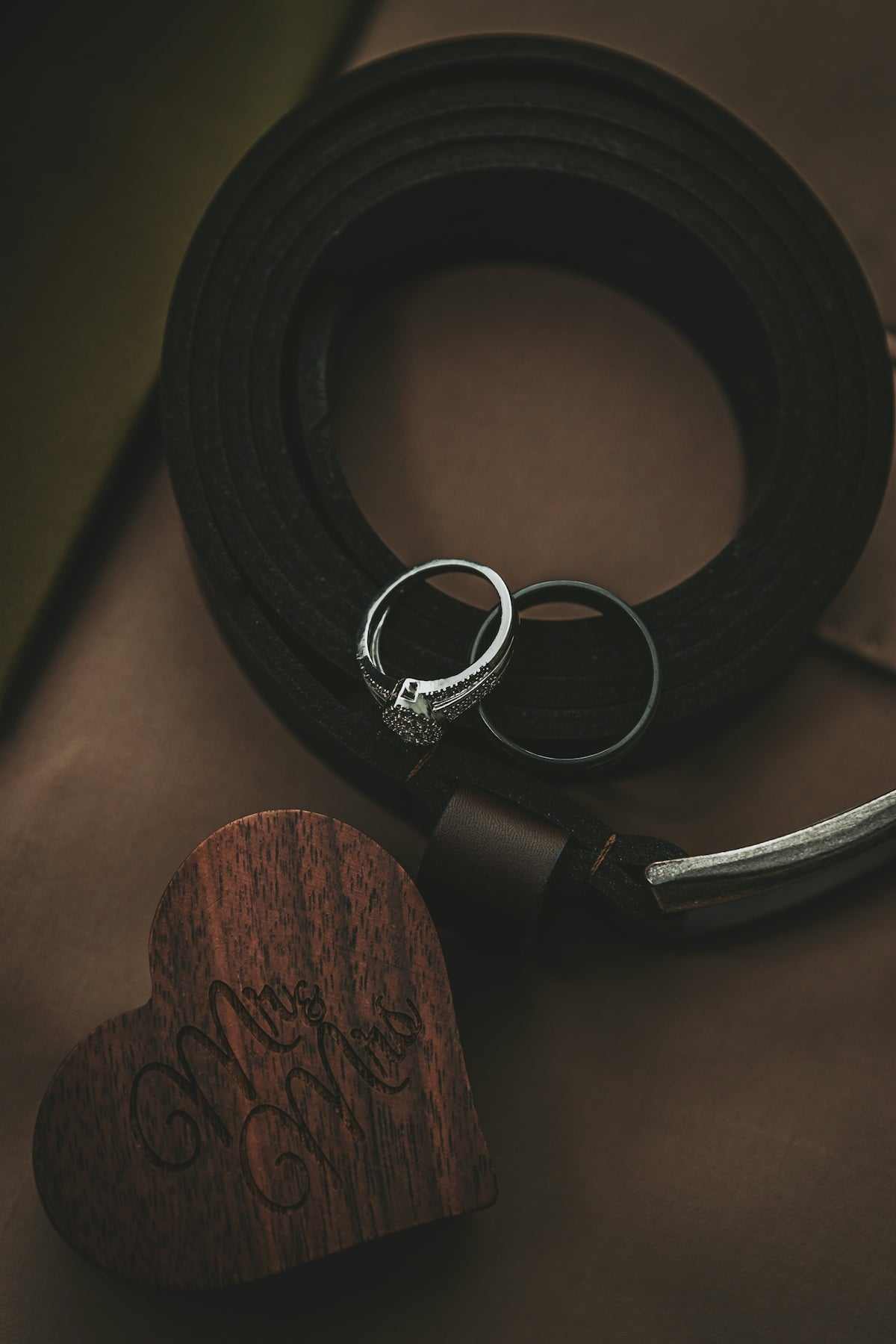 Black men's handmade leather belt with silver buckle and two rings on brown surface next to a wooden heart-shaped box